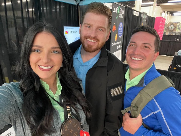 Three people are smiling indoors at an event or convention. Two men and one woman, all wearing casual attire with name tags. Behind them, booths and banners are visible. They appear to be in a lively and friendly setting.